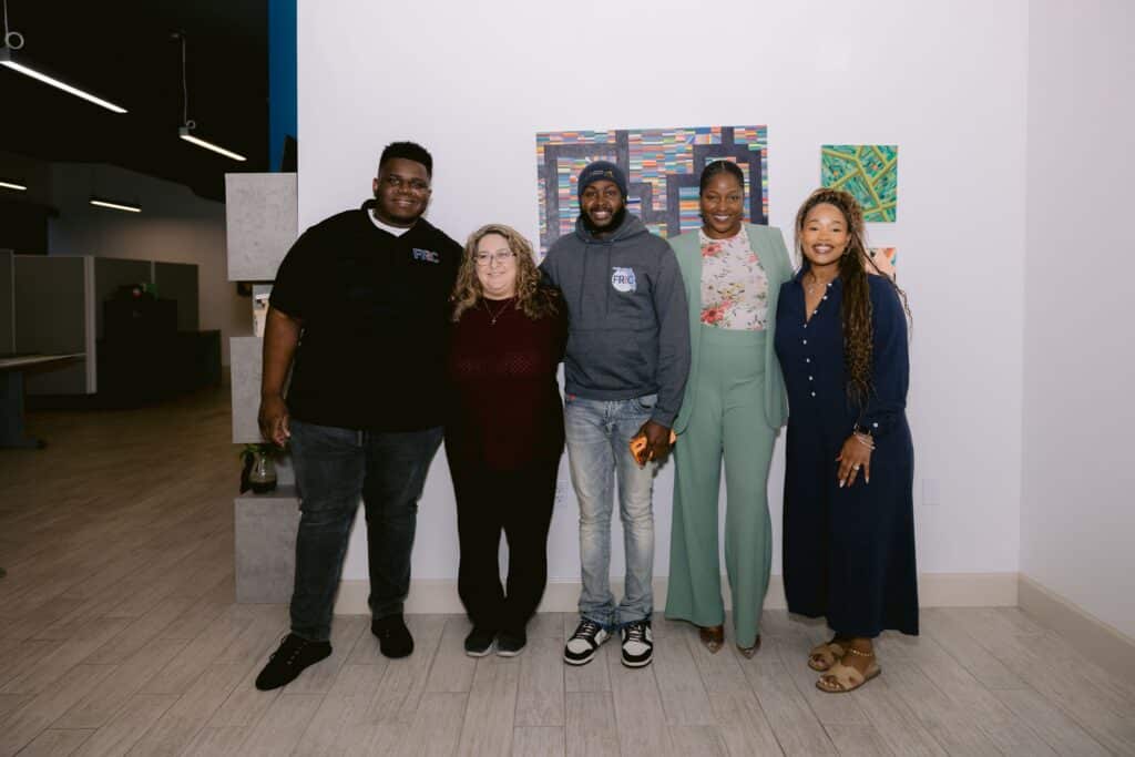 Five people of diverse backgrounds pose together indoors, smiling at the camera with colorful art on the wall behind them.