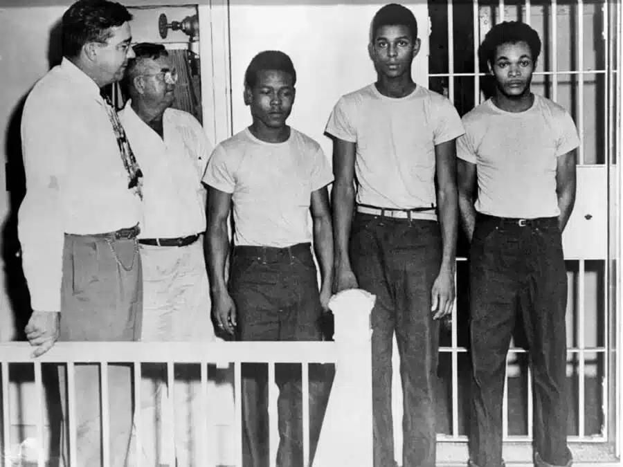 Three young Black men stand in a line in front of a barred cell, flanked by two older men in civilian clothes nearby.